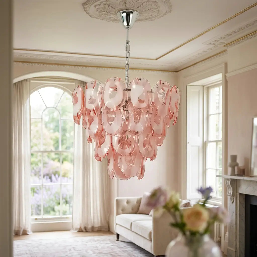 A pink glass chandelier with layered, ruffled petal-like shades hangs in a bright, elegant living room with cream walls, large windows, and a sofa in the background.